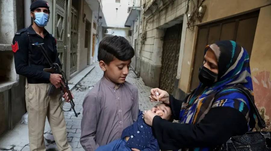 Lady health worker gives polio drops to a child with a police officer standing guard during Pakistan’s polio vaccination campaign.